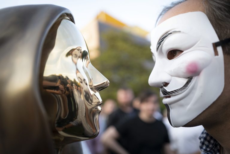 Muž v maske Guya Fawkesa pózuje so sochou anonymného tvorcu bitcoinu Satoshiho Nakamota v Budapešti. Foto: TASR/AP