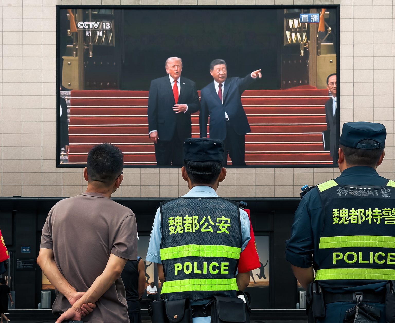 Policajti a obyvatelia pred nákupným centrom sledujú obrazovku s Xi Jinpingom a Donaldom Trumpom.